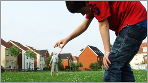 A boy wearing a red t-shirt in the foreground with another boy in the background with his hands up, he looks like he's dangling from the other boys hand.