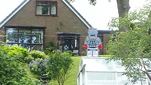 A really big robot sat on a greenhouse in the garden, using a perspective technique.