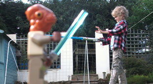 A photo of a toy close to the camera so it looks like it's sword fighting with a kid in the background