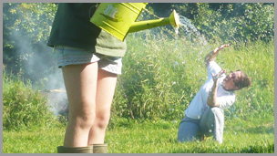 A boy in the foreground with a green watering can and a lady in the background who looks to be being watered.